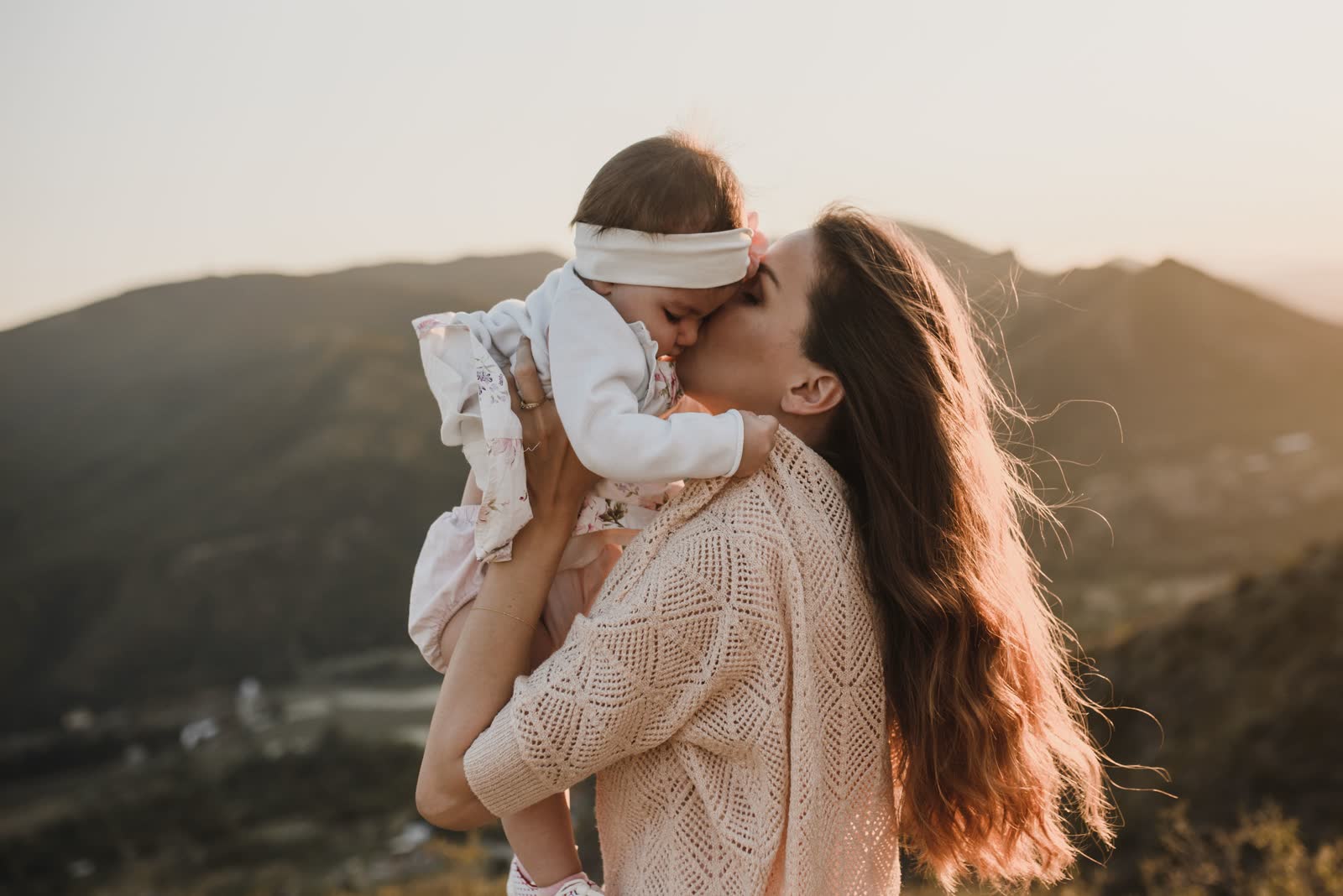 Mãe erguendo o bebê no colo e beijando seu rosto contra a luz do fim do dia, com montanhas ao fundo