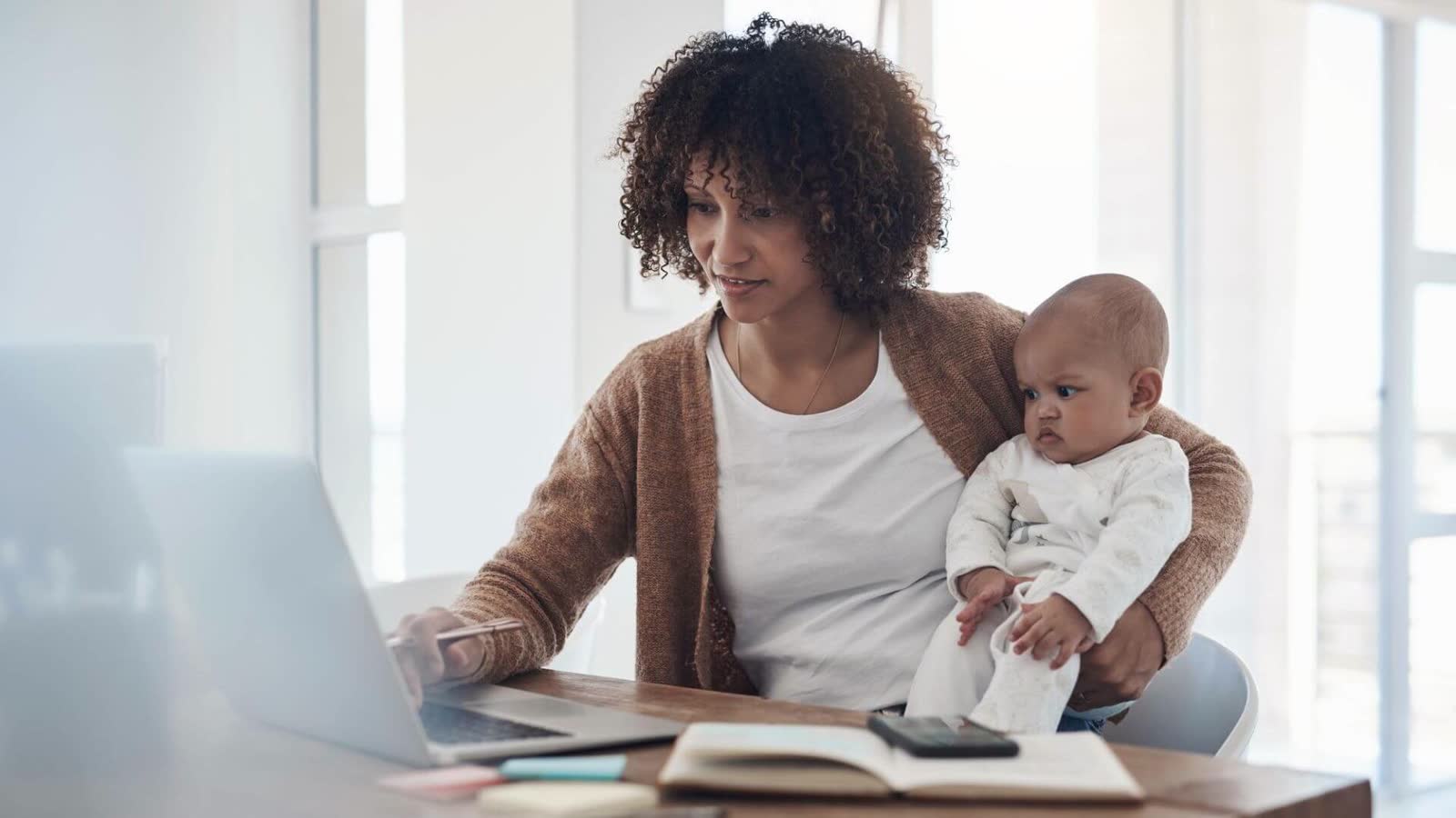 Mãe de cabelo cacheado com o bebê no colo, mexendo no notebook sobre a mesa, com caderno e celular ao lado