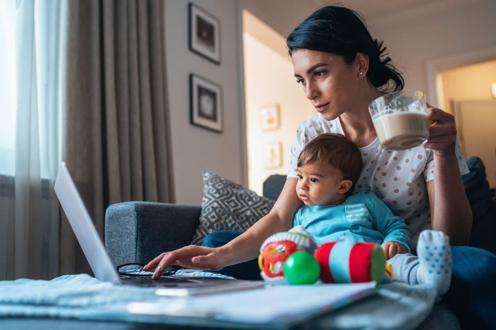 Mãe no sofá com uma xícara de café na mão, bebê no colo, mexendo no notebook em meio a papéis